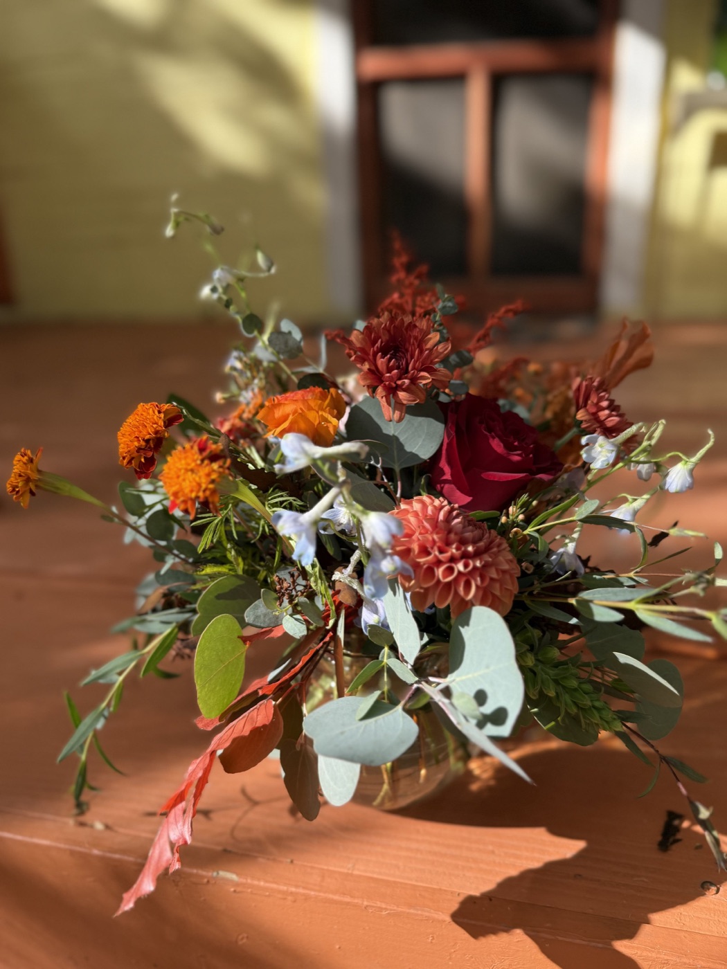 A low arrangement of rust dahlias, orange marigolds, a deep red rose and eucalyptus glowing in the afternoon sun on a porch