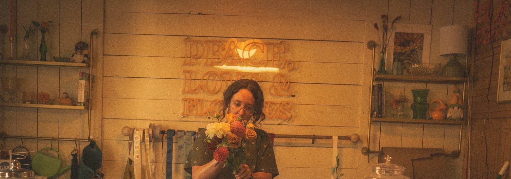 Inside Baker's Blooms: wooden shelves of vases and a glowing 'Peace, Love & Flowers' sign behind the florist's bench